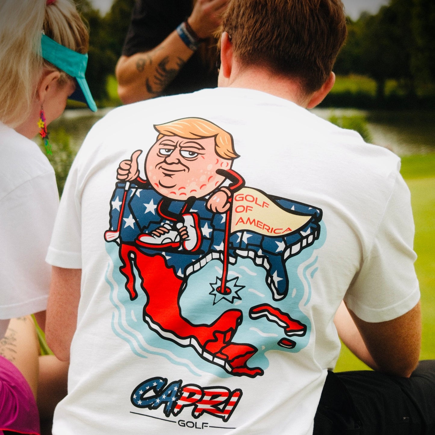 Man wearing white CAPRI-Statement T-Shirt with Golf of America caricature and stars-and-stripes backprint sitting outdoors by water
