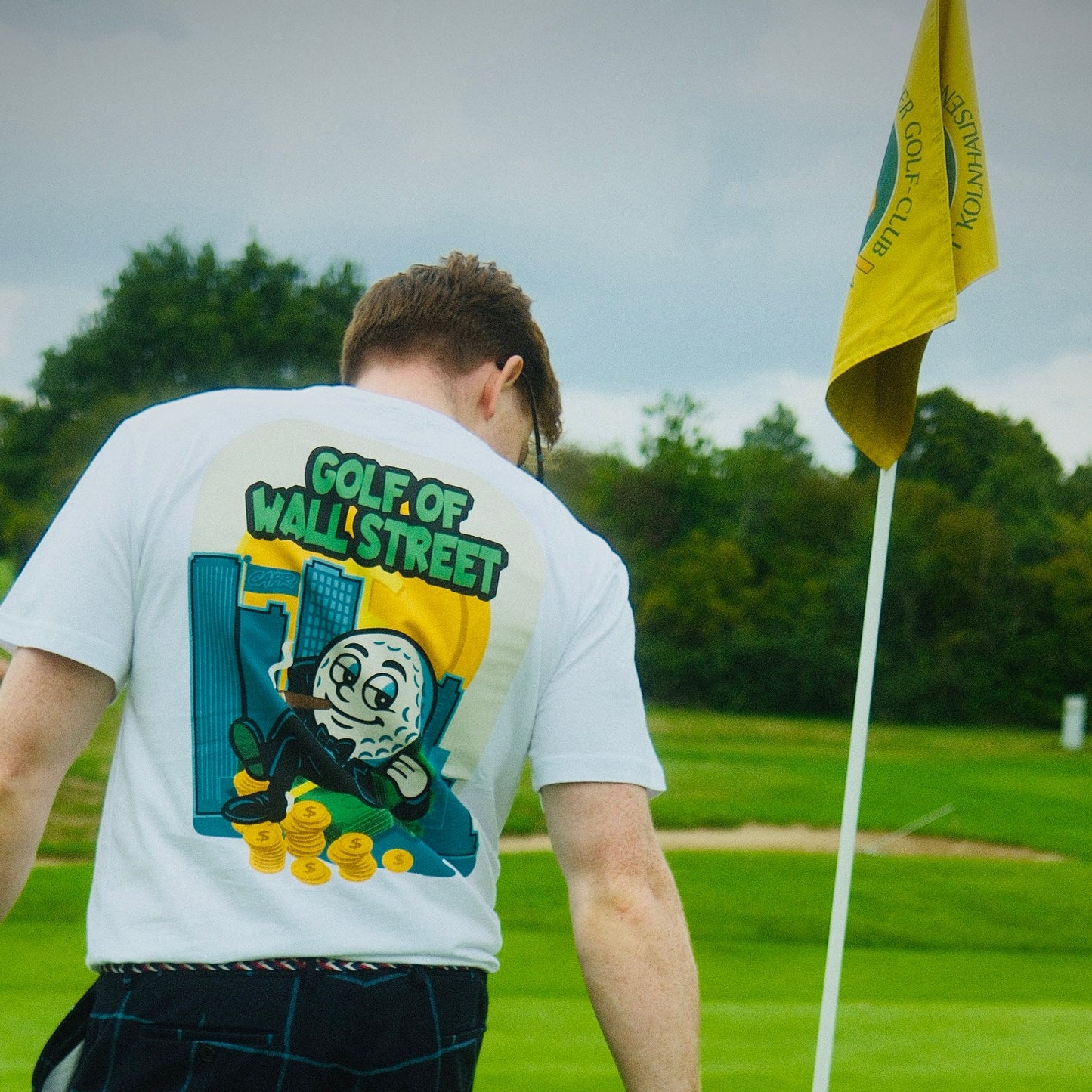 Man wearing white CAPRI-Statement T-Shirt with colorful Golf of Wall Street graphic on the back, standing on a golf course near a yellow flag.